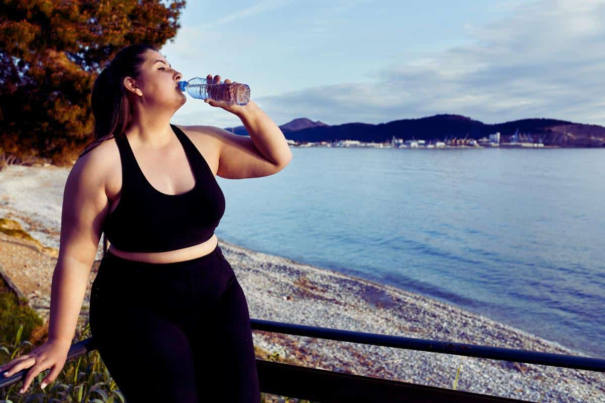 A plus size woman in black athletic wear drinks water from a bottle while sitting on a rail near a calm lakeshore. The background features trees, mountains, and partly cloudy skies, creating a serene, natural outdoor setting.