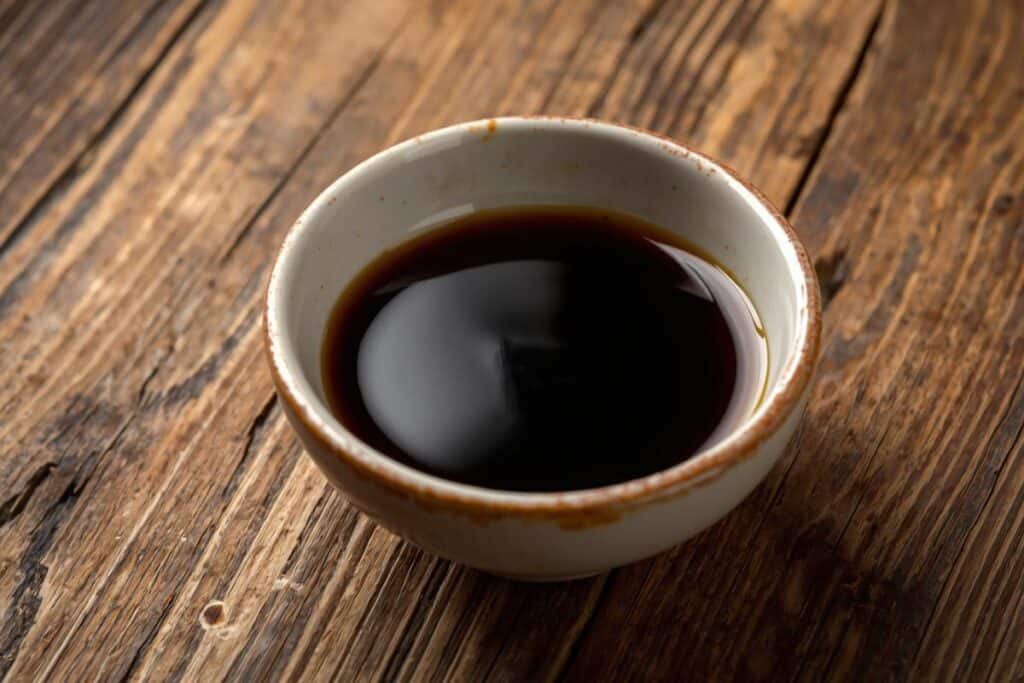 A small ceramic bowl filled with dark soy sauce sits on a rustic wooden surface.