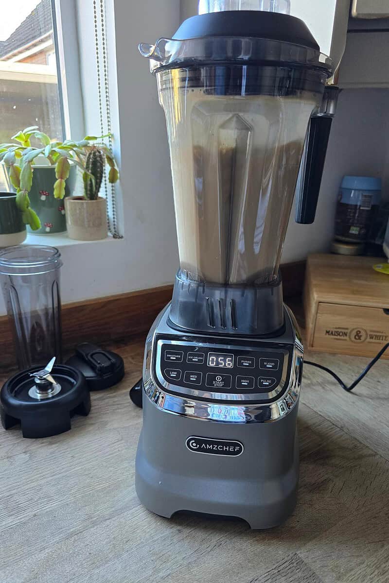 An AMZCHEF Blender sits on the kitchen counter, diligently mixing a light brown concoction. The digital timer shows 56 seconds remaining. Nearby, a plant and wooden container add warmth to this culinary scene, perfect for your next kitchen review of the Model 5003.