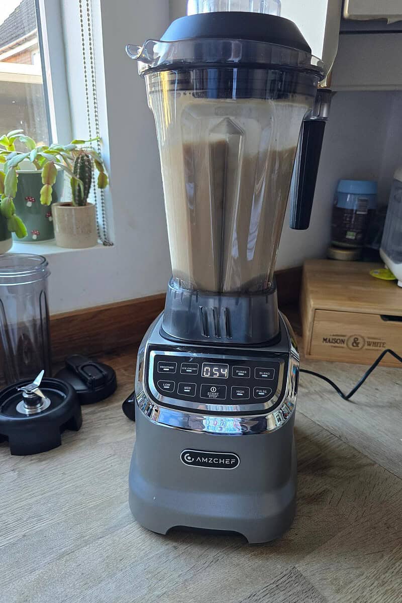 An AMZCHEF blender with a digital display and control panel blends a light brown liquid on a kitchen counter near a window with potted plants. The blender lid, a cup, and a wooden container labeled “Mason & White” sit nearby.