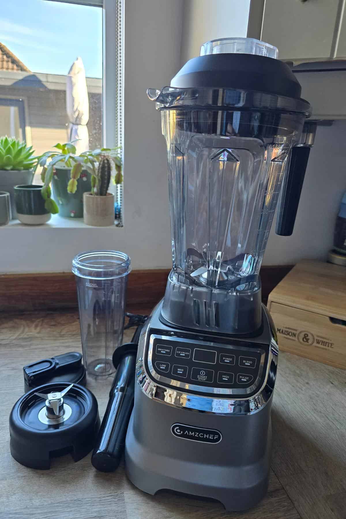 The AMZCHEF Blender, Model 5003, features a clear pitcher on a kitchen counter and comes with a separate blade attachment, tamper, and personal blender cup. Potted plants adorn the windowsill in the background, setting a perfect scene for your kitchen review.
