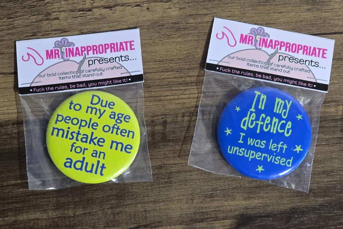 Two packaged novelty badges rest on a wooden surface from the Rude Gift Website. The left one says, "Due to my age, people often mistake me for an adult." Meanwhile, the right quips, "In my defense, I was left unsupervised." For those moments that make you laugh and blush!.