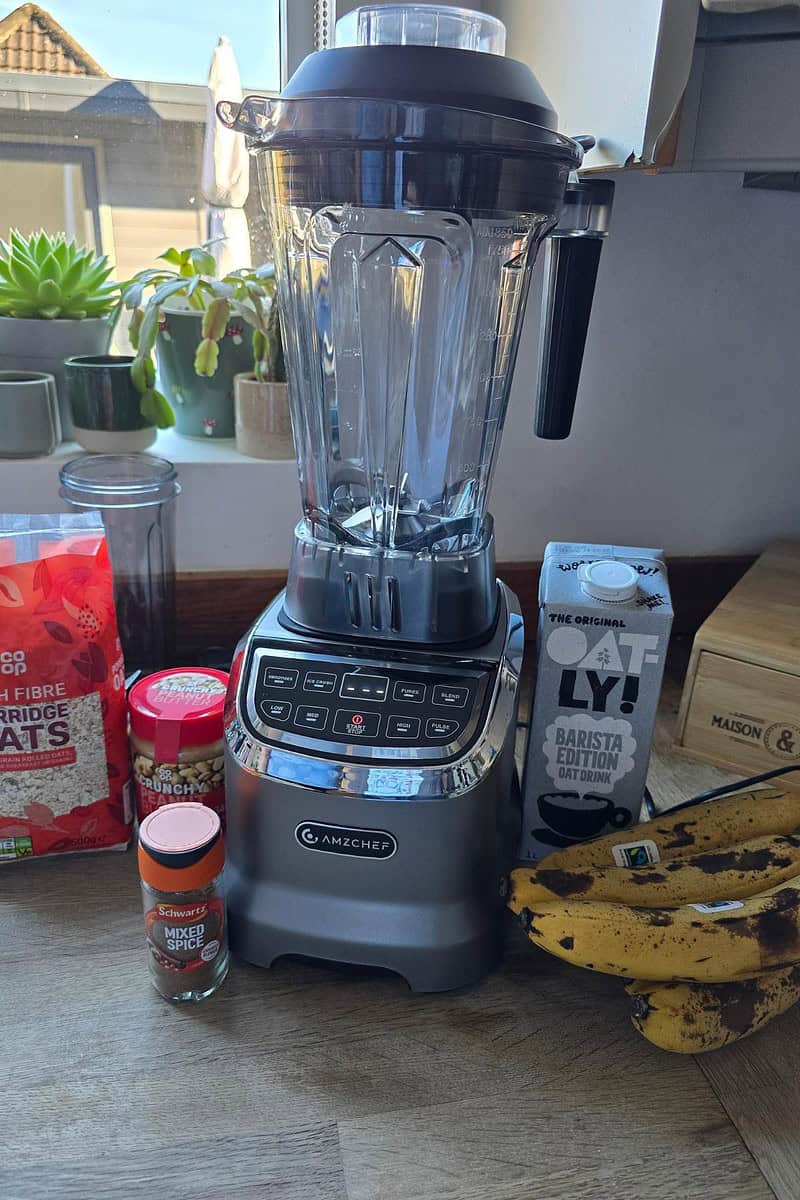 The AMZCHEF Blender, Model 5003, sits proudly on the kitchen counter, surrounded by a juice bottle, porridge oats, mixed spice container, oat milk, and ripe bananas.