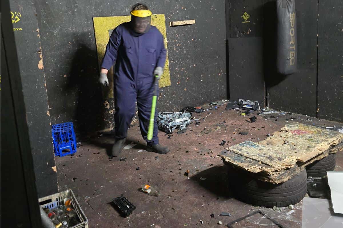 In a chaotic room filled with debris and a punching bag, a person in a blue jumpsuit and yellow face shield wields a green tool, ready to unleash mayhem: welcome to the world of Smash or Miss, where every visit is an exhilarating review of pent-up fury at Destroy'd Rage Rooms.