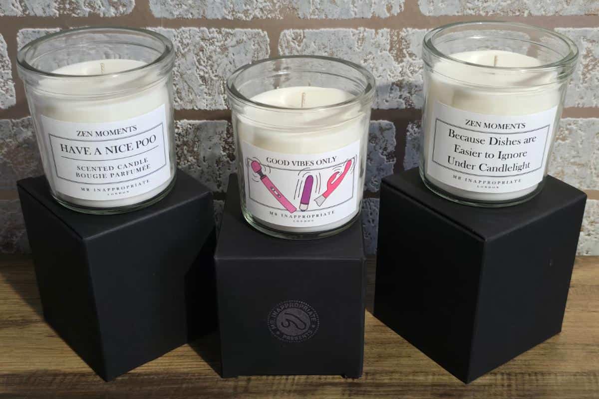 Three glass jar candles sit atop black boxes, perfect for a Mr Inappropriate Review. The left reads "HAVe A NICE POO," the middle "GOOD VIBES ONLY" with pink hands, and the right "Because Dishes are Easier to Ignore Under Candlelight." All are ZEN MOMENTS.