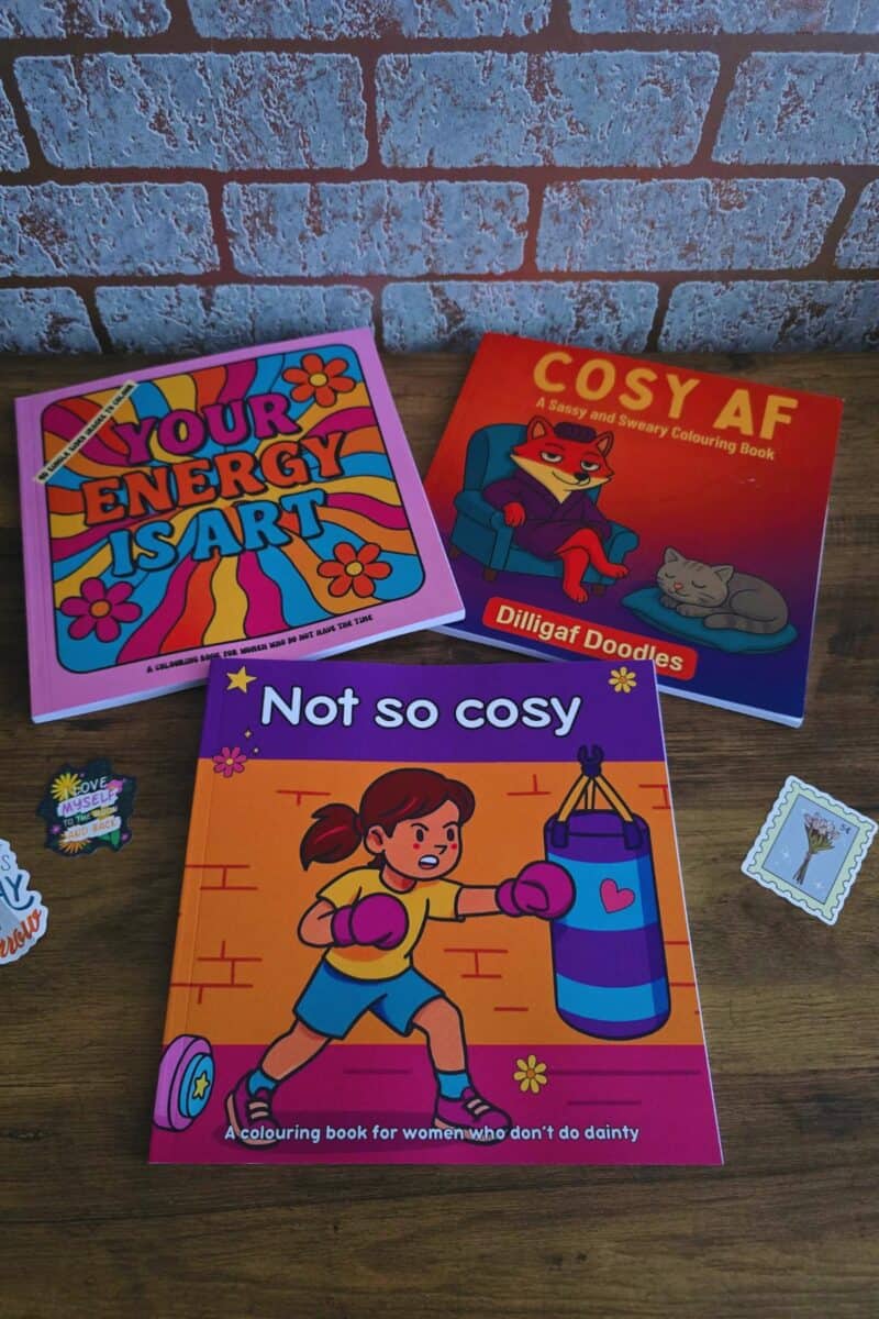 Three adult colouring books are displayed on a wooden surface: “Your Energy Is Art” with a vibrant, retro design, “Cosy AF” featuring a cat illustration, and “Not So Cosy” showing a cartoon woman boxing. Stickers are also visible.