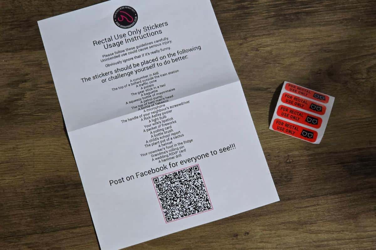 A paper titled "Rectal Use Only Stickers Usage Instructions" with a QR code lies beside cheeky "For Rectal Use Only" stickers on a wooden surface. Perfect for those who enjoy a laugh and blush, find them on Rude Gift Website or see how they fare in a Mr. Inappropriate Review!.