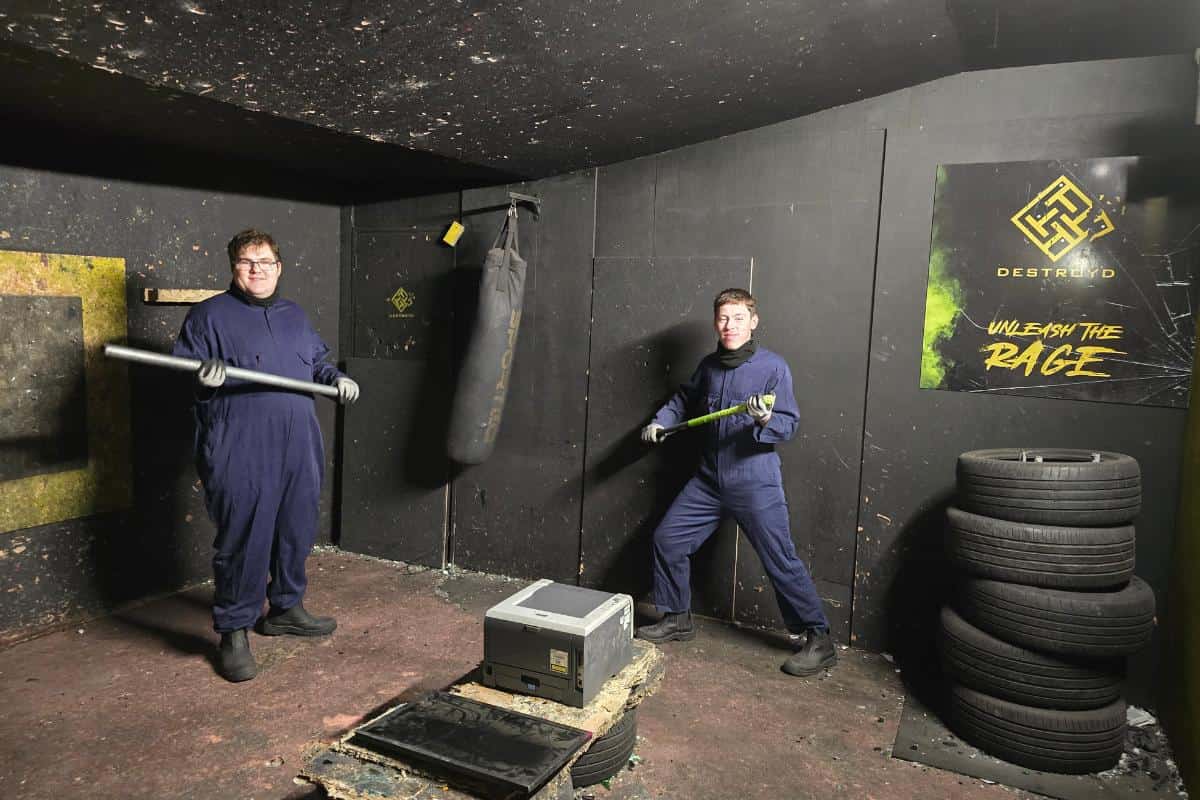 Two people in protective suits hold bats, ready to smash their stress away in a rage room with a punching bag and stacked tires.