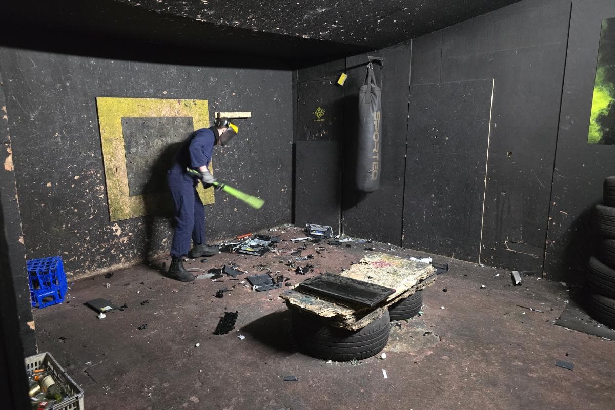 In a wild rage room, a person in protective gear fiercely swings a bat, smashing objects amidst debris, tires, and a punching bag.