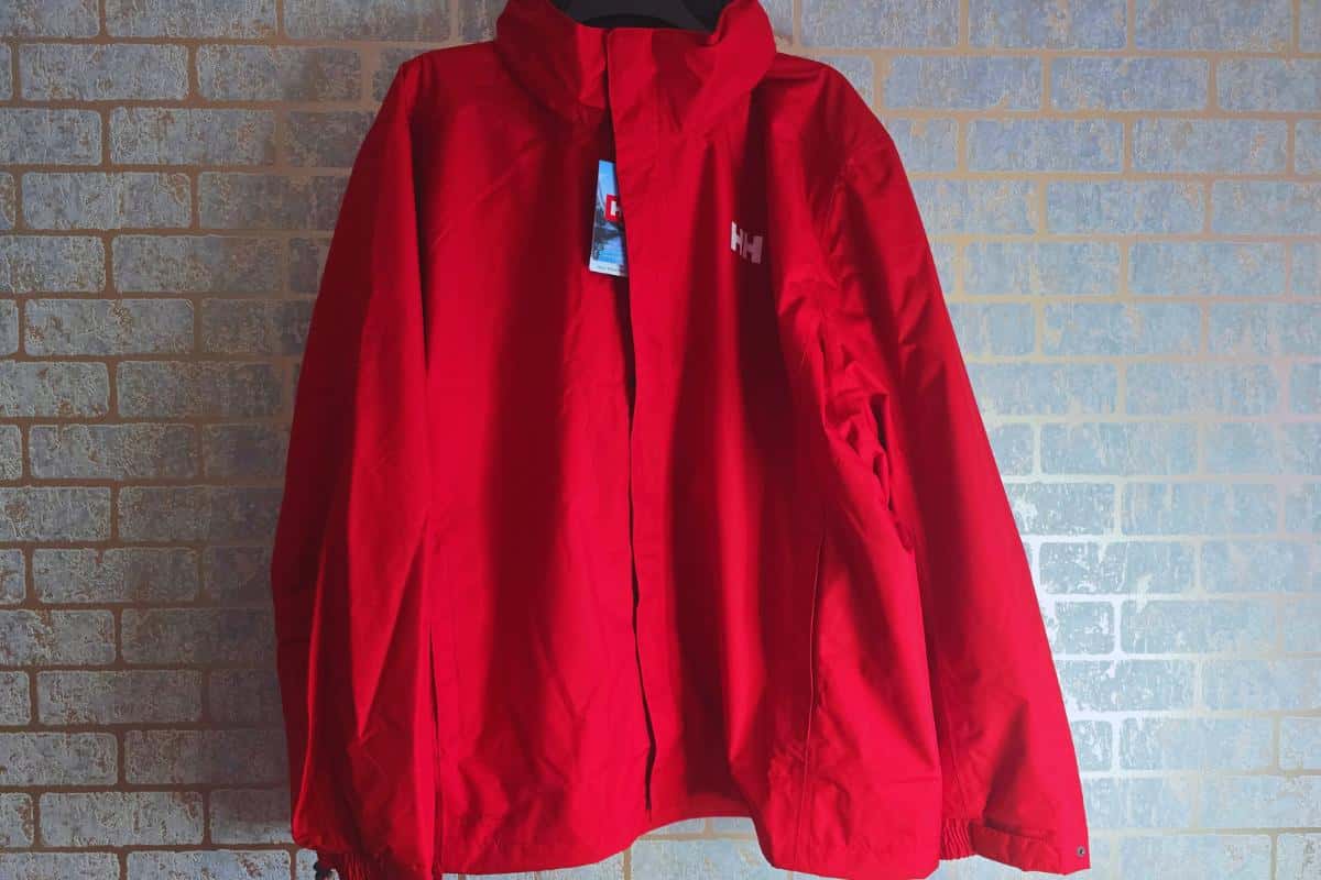 A bright red Helly Hansen jacket, ideal for plus-size hiking, hangs against a light-colored brick wall. It features a high collar, long sleeves, front zipper, visible chest logo, and a blue tag near the collar.