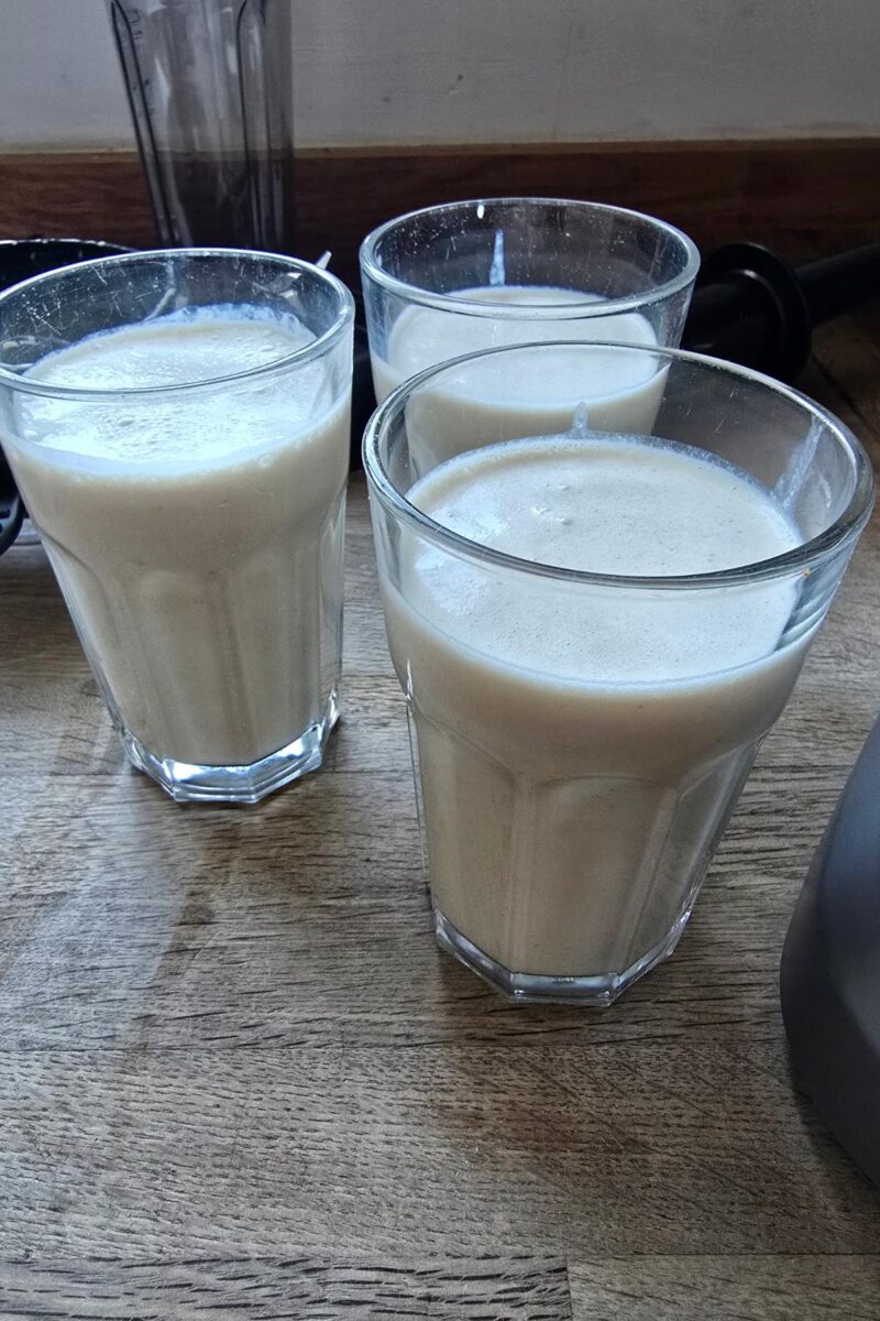 Three clear glasses filled with a creamy light-colored beverage are placed on a wooden surface, showcasing the smooth results achieved by the AMZCHEF Blender. Perfectly blended, this kitchen marvel, Model 5003, ensures every sip is as delightful as the last.