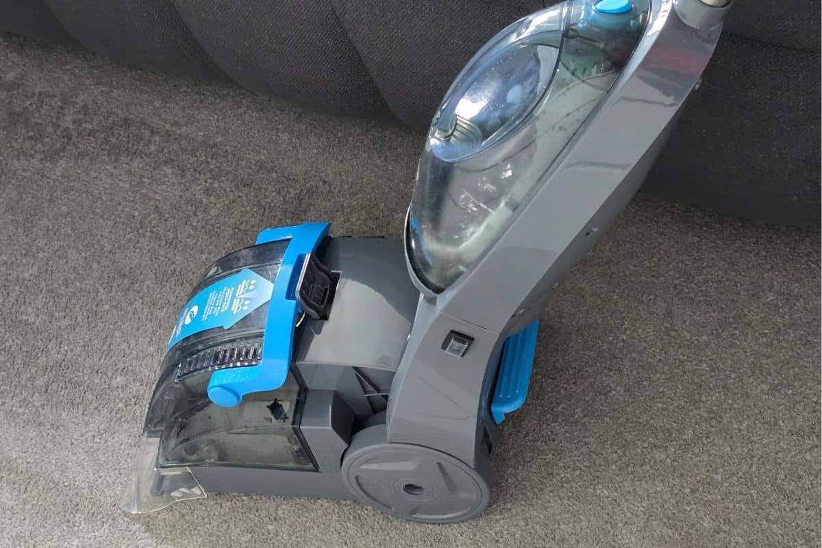 The AquaJetPlus gray and blue carpet washer stands ready on the carpeted floor, poised in front of a dark upholstered sofa.