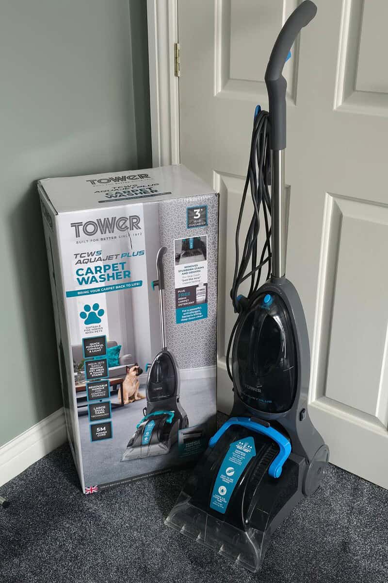 The TCW5 AquaJetPlus carpet washer is stationed elegantly beside its packaging box in a room adorned with a gray carpet and green walls, inviting you to explore its remarkable cleaning features.