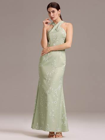 A person stands against a neutral background wearing a long, sleeveless, light green dress with a high neckline and floral pattern. The hair is tied back, and the person is wearing earrings and bracelets.