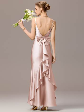 A woman in a light pink, backless evening gown with a bow and ruffle detail stands holding a bouquet of white flowers. Her hair is styled in an updo, and she is wearing high heels. The background is a neutral color.
