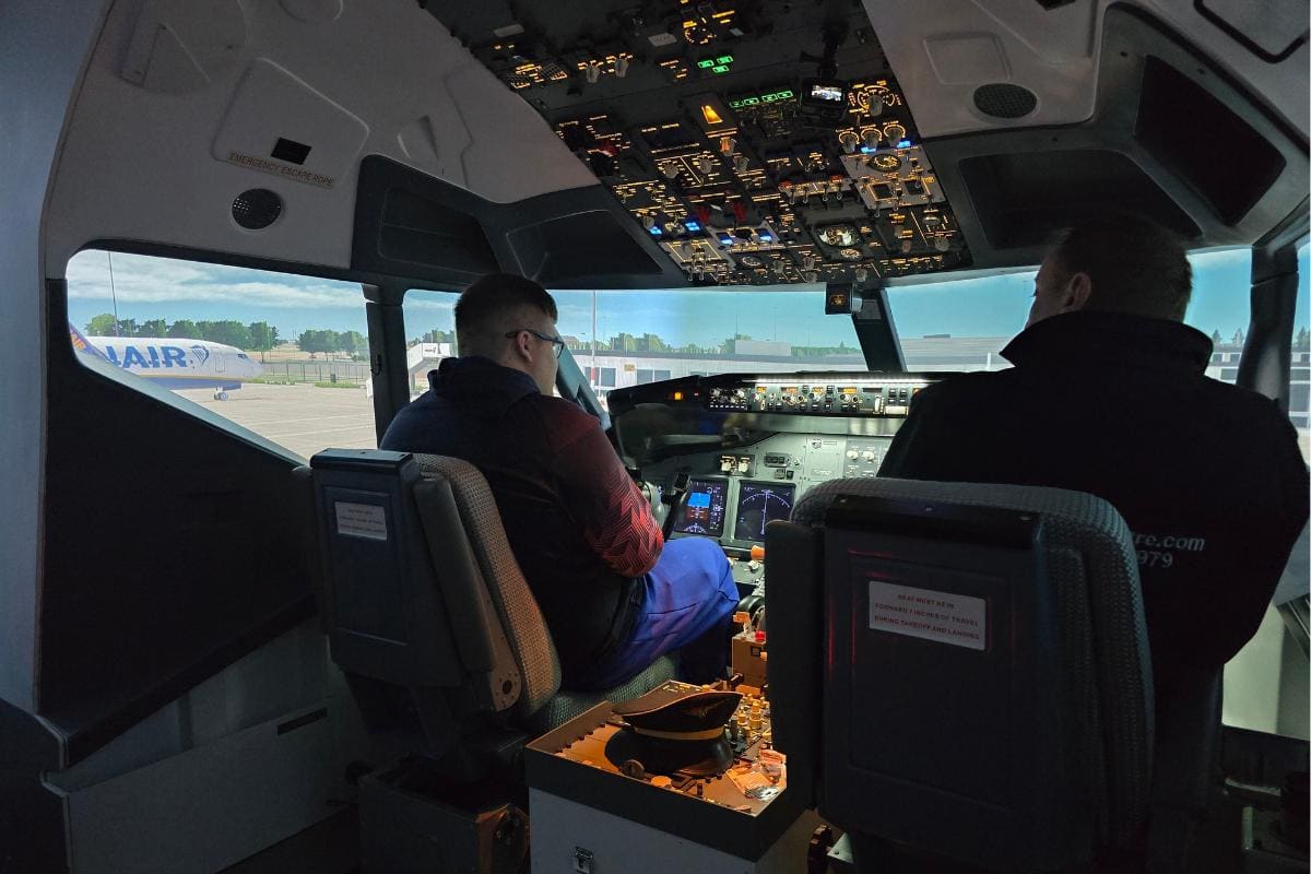 Two individuals are seated in a FlightSimCentre cockpit, surrounded by control panels and displays. Outside, an aircraft is visible through the window on the left, creating a realistic flight simulator experience that captures the essence of an actual airport scene.