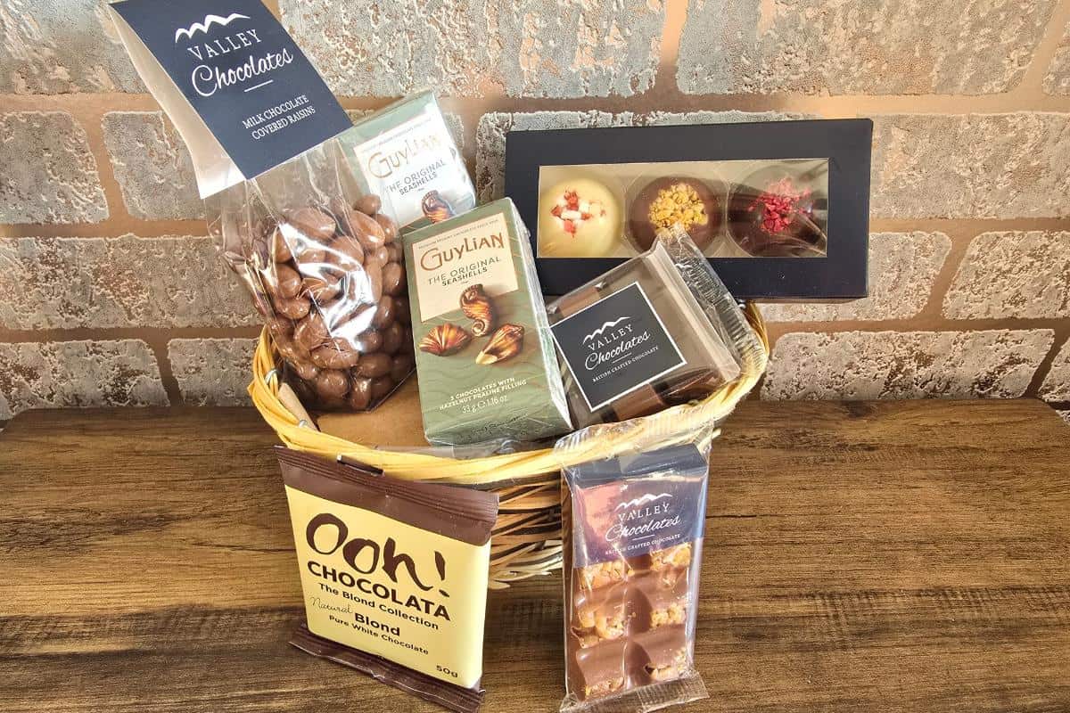 A wicker basket on a wooden surface contains assorted chocolates, including chocolate-coated nuts, Guylian chocolate bars, boxed truffles, a chocolate square, and a blond chocolate drink sachet. The background is a light brick wall.