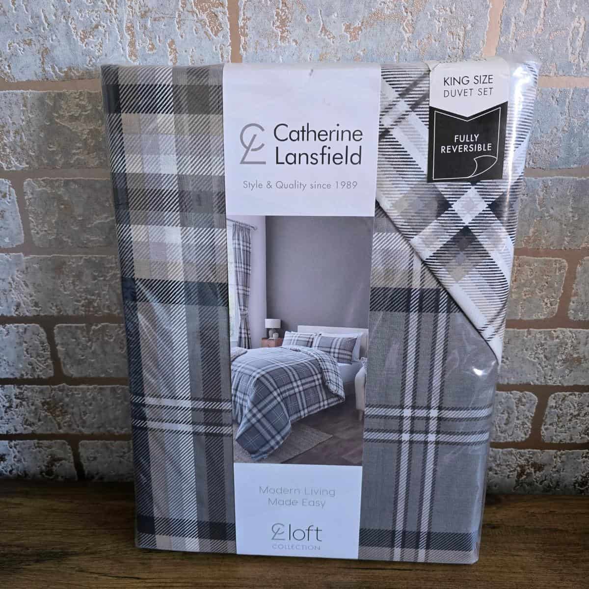 A packaged Catherine Lansfield king size duvet set, featuring a grey, white, and black plaid pattern. The packaging shows a bedroom with the duvet on a bed. Labels indicate it is fully reversible and part of the Loft Collection. Background: brick wall.