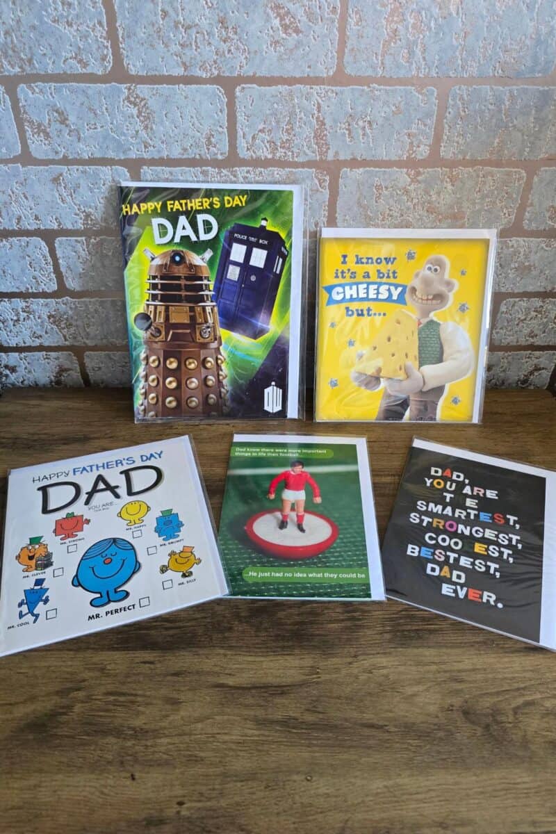Five Father’s Day cards on a wooden surface: One with Doctor Who characters, one with Wallace and Gromit holding cheese, one with Mr. Men drawings, one showing a football toy, and one with bold white text listing positive traits, all against a light brick wall.