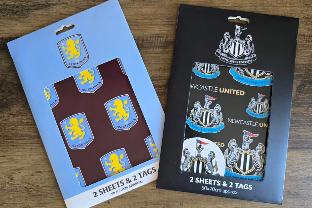Two packs of football-themed gift wrap lie on a wooden surface. The left pack is blue with Aston Villa FC crests, while the right pack is black with Newcastle United FC crests and gold “NEWCASTLE UNITED” text. Both packs contain 2 sheets and 2 tags.