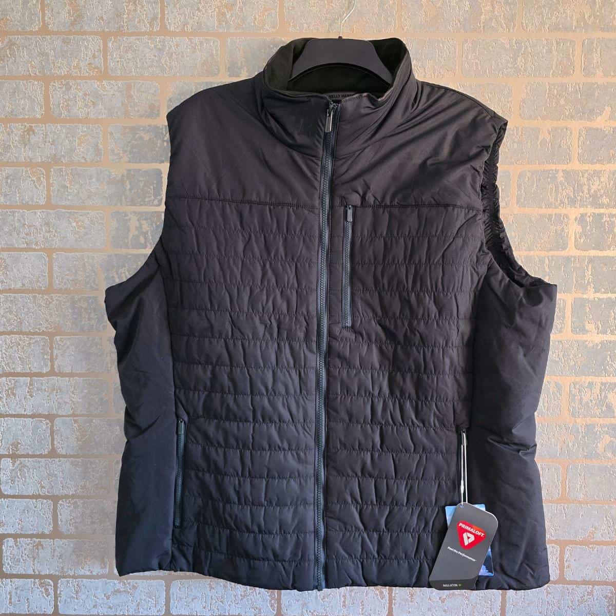 A black sleeveless puffer vest hangs on a hanger against a light brown brick wall. The vest features a quilted front, zippered chest pocket, side pockets with zippers, and a tag attached near the bottom hem on the right side.