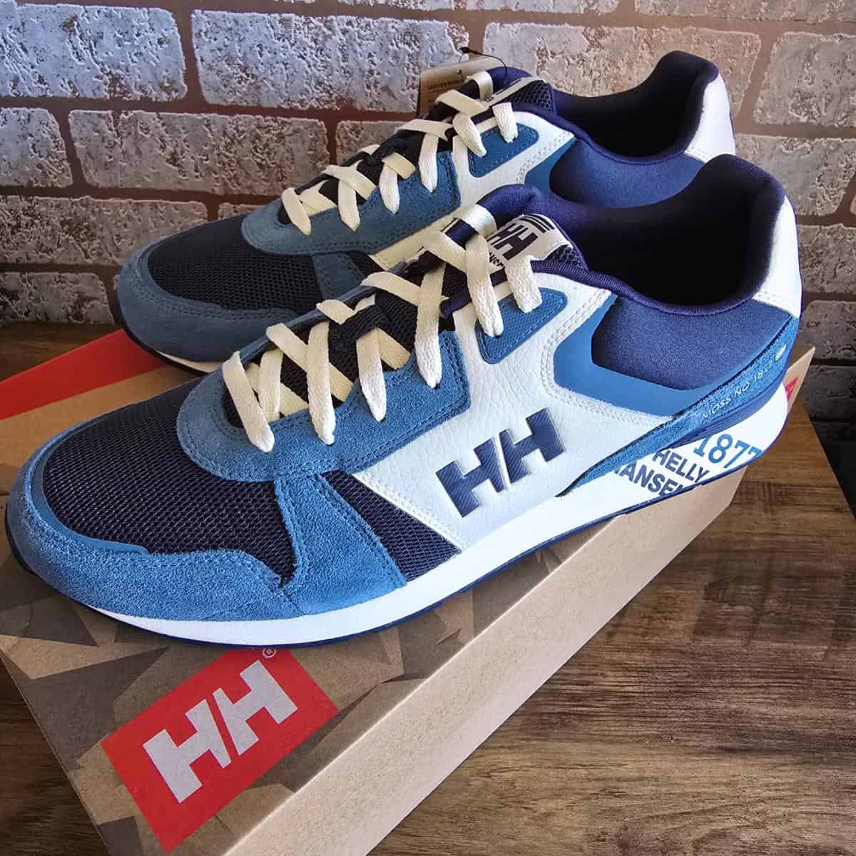 A pair of blue, white, and black Helly Hansen sneakers with cream laces sits on top of a brown Helly Hansen shoebox. The shoes feature the HH logo on the side and branding near the heel. The background is a rustic brick wall and wooden surface.