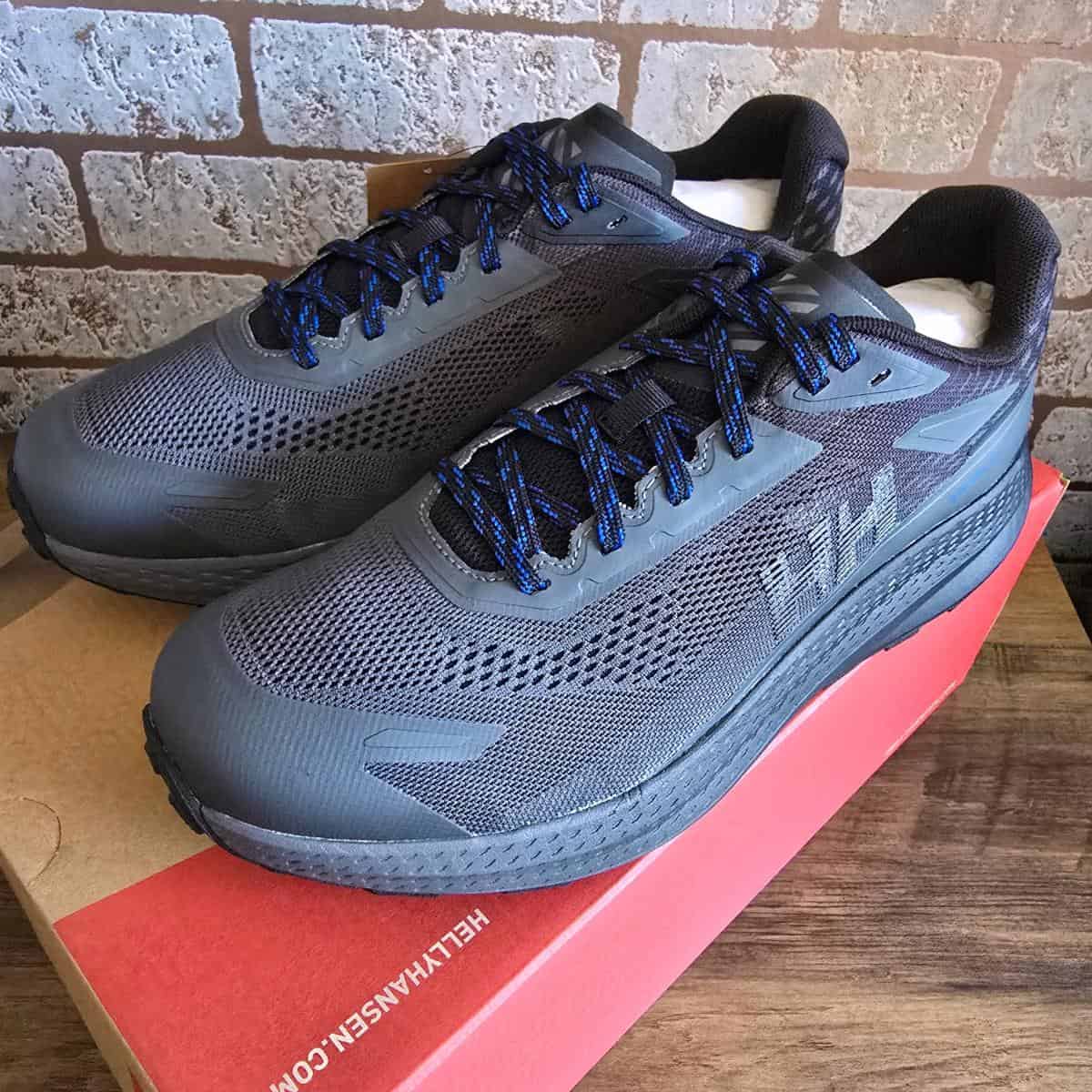 A pair of dark gray athletic sneakers with blue and black laces sits on top of a red and brown Helly Hansen shoebox. The shoes have mesh uppers, textured soles, and “HH” branding on the side. The background is a brown brick wall.