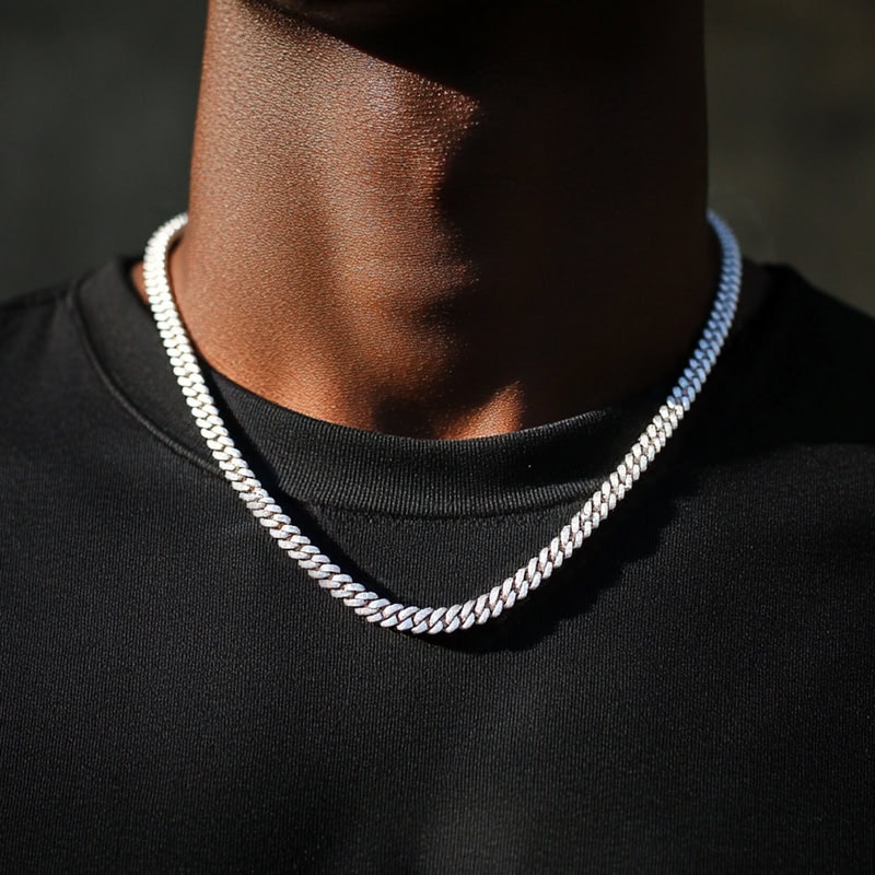 A close-up of a person wearing a plain black t-shirt and a thick silver Cuban link chain necklace. The focus is on the men's jewellery, with the blurred dark background highlighting the necklace and fabric texture.