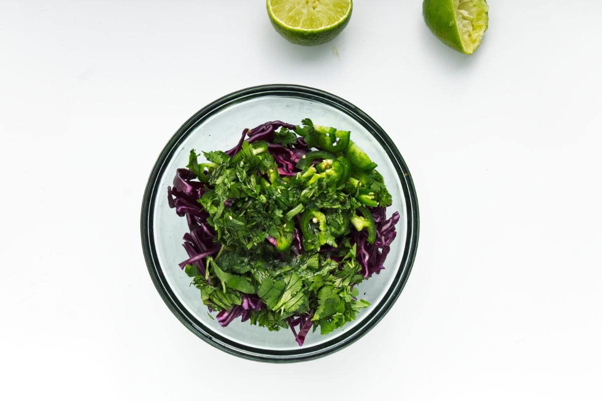 A glass bowl filled with Spicy Cabbage Slaw—shredded purple cabbage, chopped cilantro, and sliced green jalapeño—sits on a white surface. Perfect for topping Roasted Cauliflower Tacos or as an easy dinner side, the ingredients appear fresh and vibrant.
