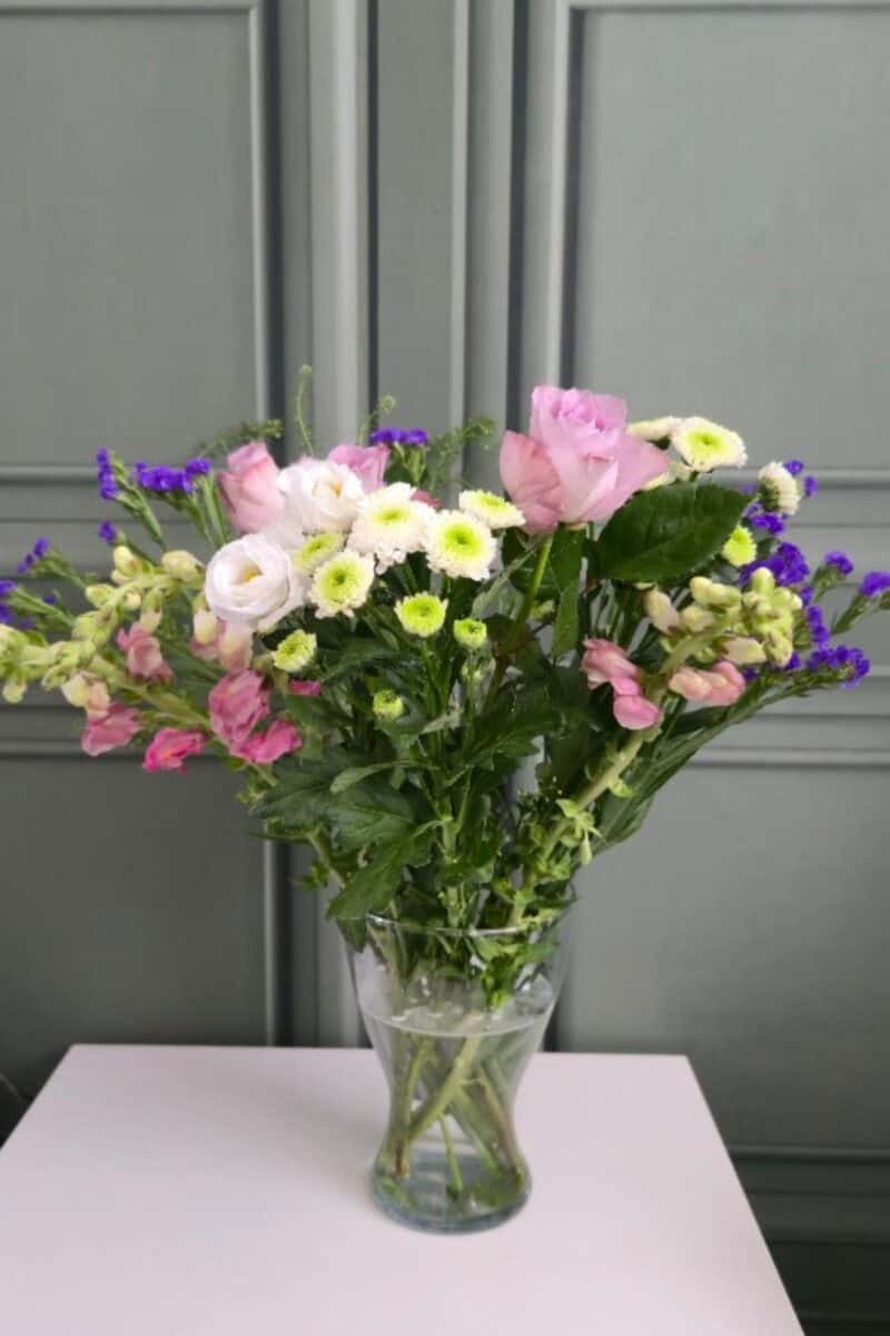 A clear glass vase from Prestige Flowers holds a mixed bouquet of pink roses, white daisies, light pink ranunculus, and purple statice flowers. Displayed on a pale surface with a gray paneled background, the arrangement looks fresh and elegant year-long.