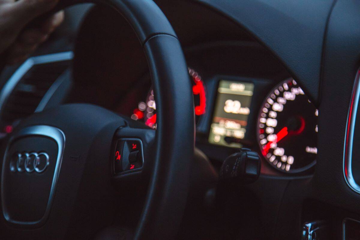 Close-up of an Audi steering wheel and dashboard, showing illuminated speedometer, tachometer, and infotainment screen with red lighting. A hand grips the wheel—perfect for your car insurance checklist before that summer coverage road trip.