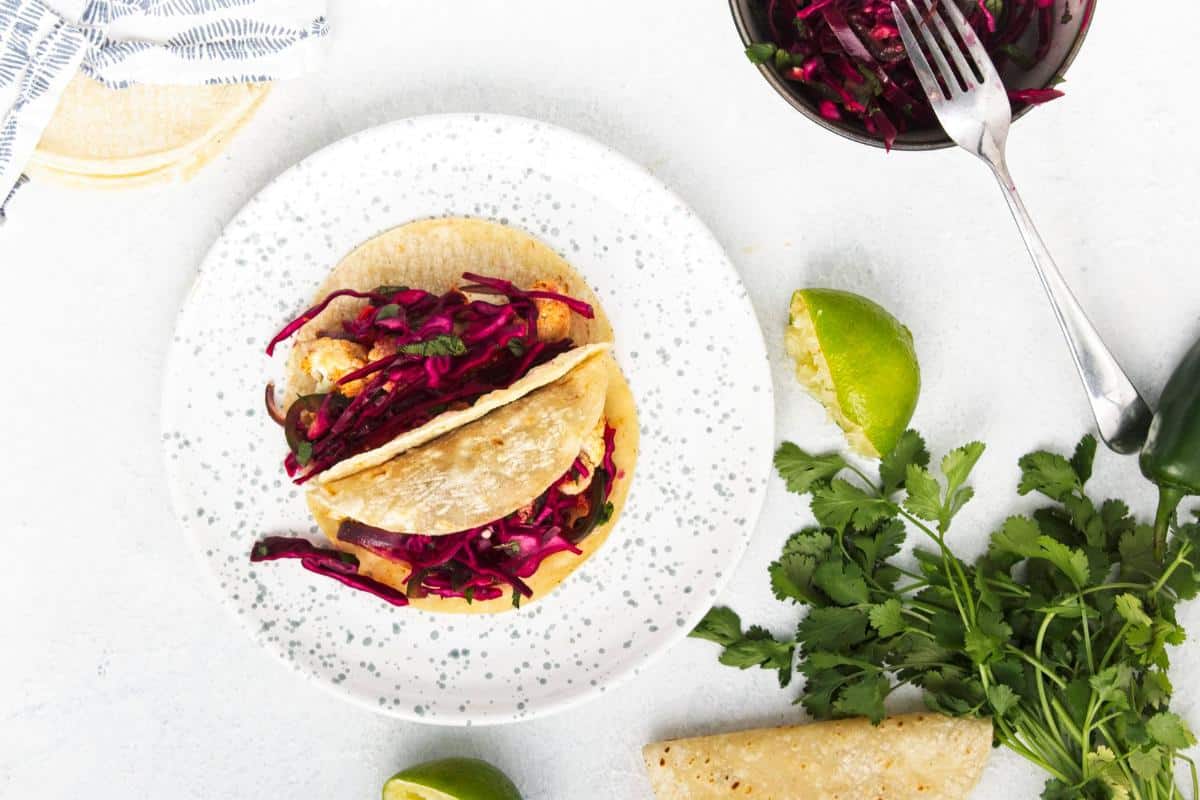 A plate with two Roasted Cauliflower Tacos filled with spicy cabbage slaw sits on a white surface. Nearby are cilantro, lime wedges, a partial tortilla, and a bowl of slaw with a fork—an easy dinner scene. A striped napkin is in the top left corner.