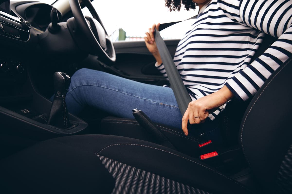 A person wearing a striped shirt and jeans sits in a car and fastens their seatbelt. The photo, taken from the side, highlights safe driving—an important step before hitting the road with your car insurance in place.