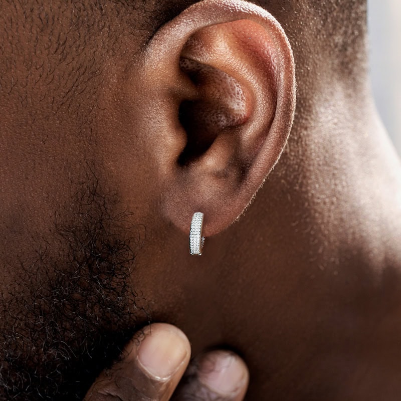 Close-up of a person’s ear wearing a small, silver, diamond-studded hoop earring—a stylish piece of men's jewellery. The person's dark skin and beard are visible as their hand gently touches their neck, focusing on the earring's formal jewellery details.