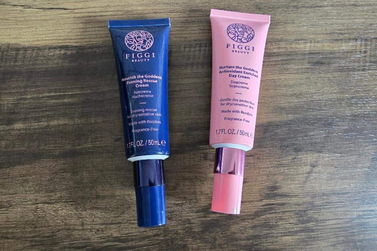 Get a first look at FIGGI Beauty Products: two skincare tubes on a wooden surface. The dark blue “Nourish the Goddess Revitalizing Rescue Cream” sits beside the pink “Nurture the Goddess Antioxidant Day Cream.”.