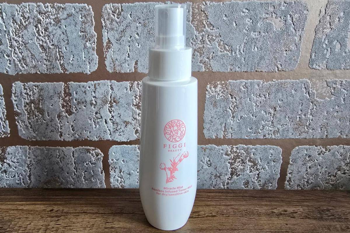 Get a first look at a white FIGGI BEAUTY spray bottle with a pink logo and text, standing on a wooden surface against a light brown and gray brick wall—perfect for your holiday Figgi Beauty Products collection.