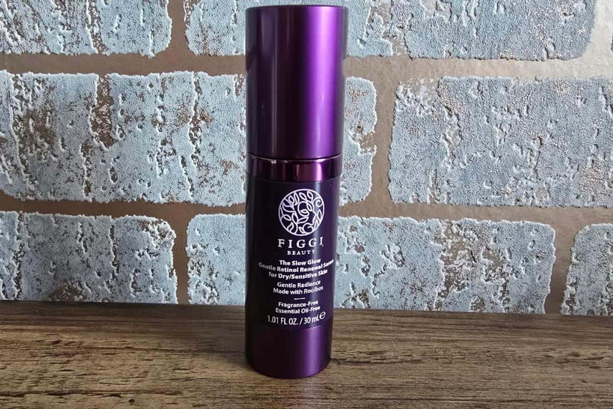 A purple bottle of Figgi Beauty Products stands on a wooden surface in front of a brick-patterned background, offering a festive first look at this holiday skincare essential. The label features a white tree logo and product details in white text.