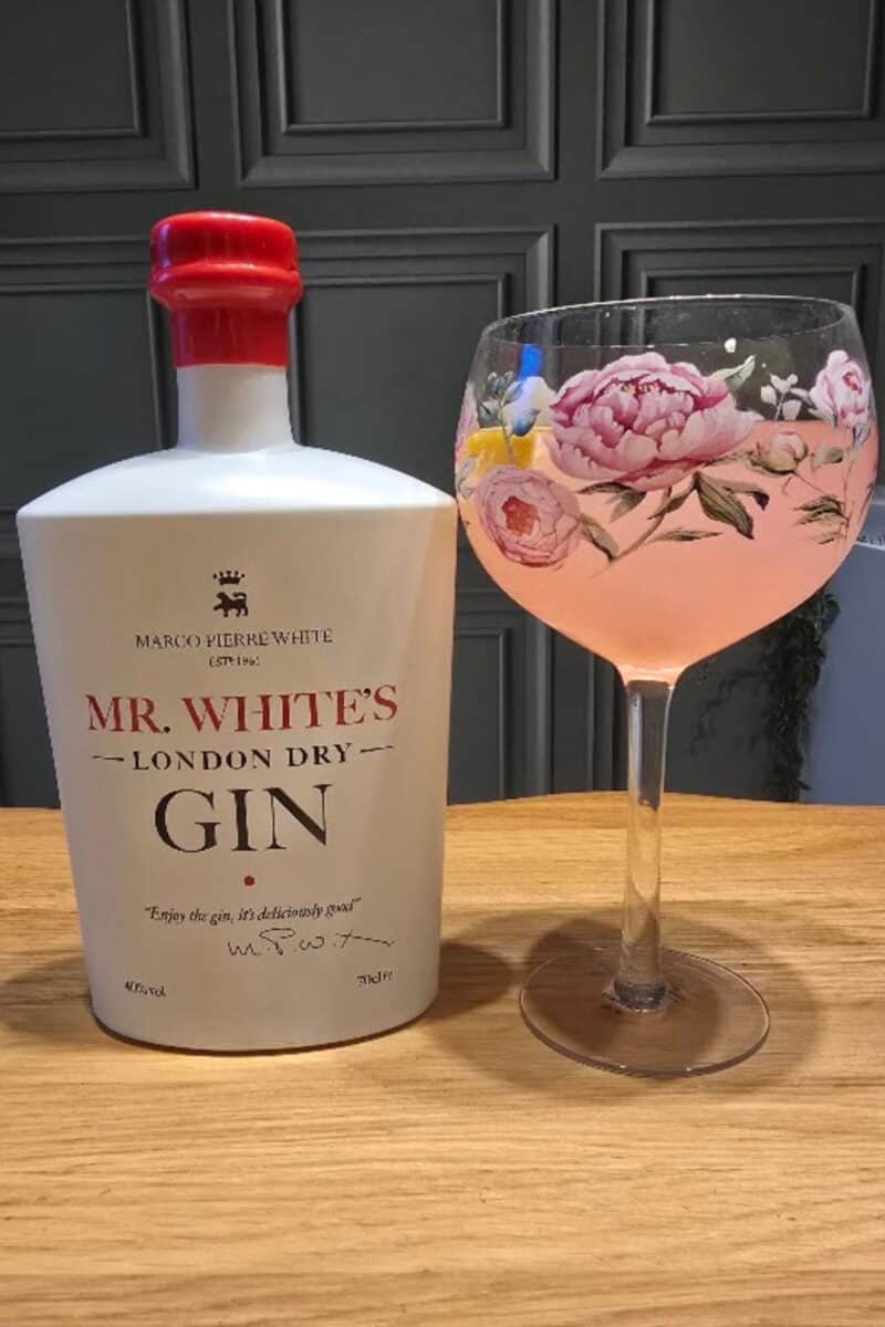 A white bottle of Mr White's Gin with a red cap stands beside a large glass of pink cocktail adorned with floral patterns, lemon, and a flower garnish, all set on a wooden surface—an inviting setup for any taste test.