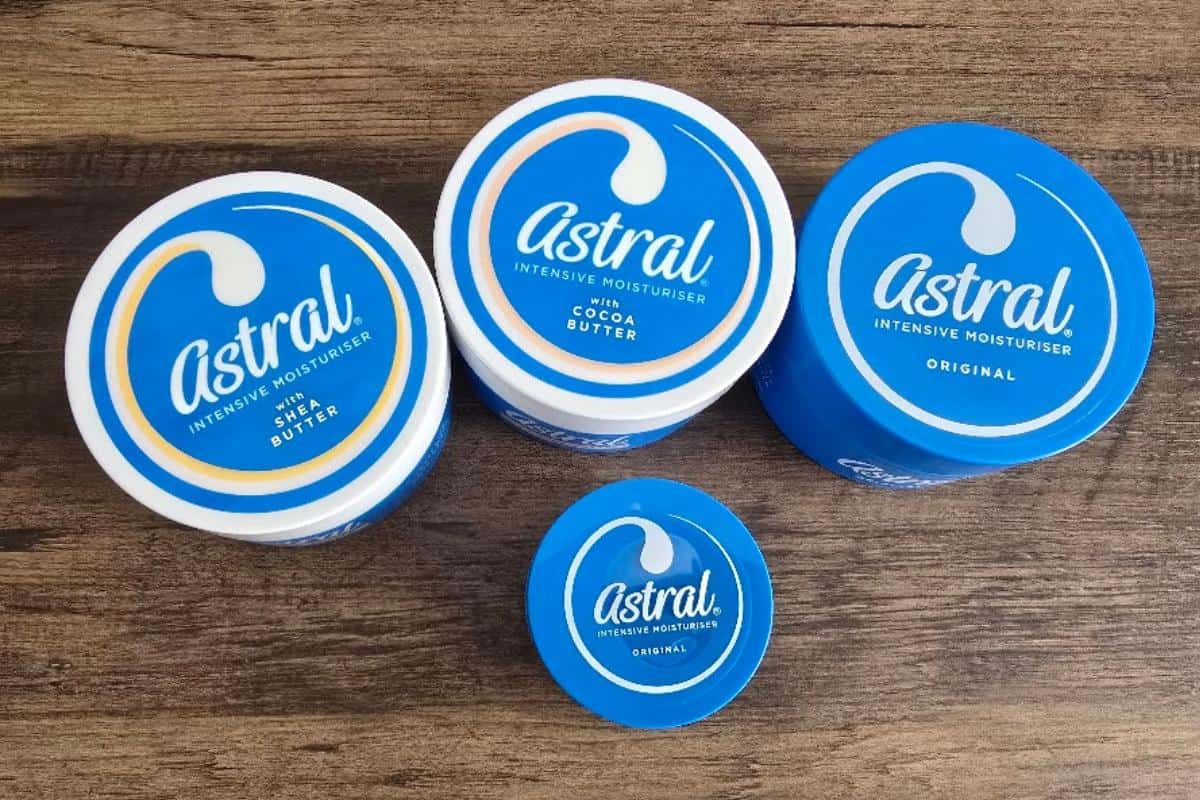 Four blue tubs of Astral Intensive Moisturiser in different sizes, arranged on a wooden surface—perfect European Road Trip essentials. Two tubs are labeled Original, one Cocoa Butter, and one Shea Butter.
