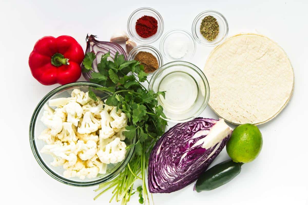 A flat lay of fresh ingredients on a white surface for Easy Dinner Roasted Cauliflower Tacos: red bell pepper, half a red onion, garlic cloves, jalapeño, lime, cilantro, cauliflower florets in a bowl, tortillas, oil, and spices—perfect with Spicy Cabbage Slaw.