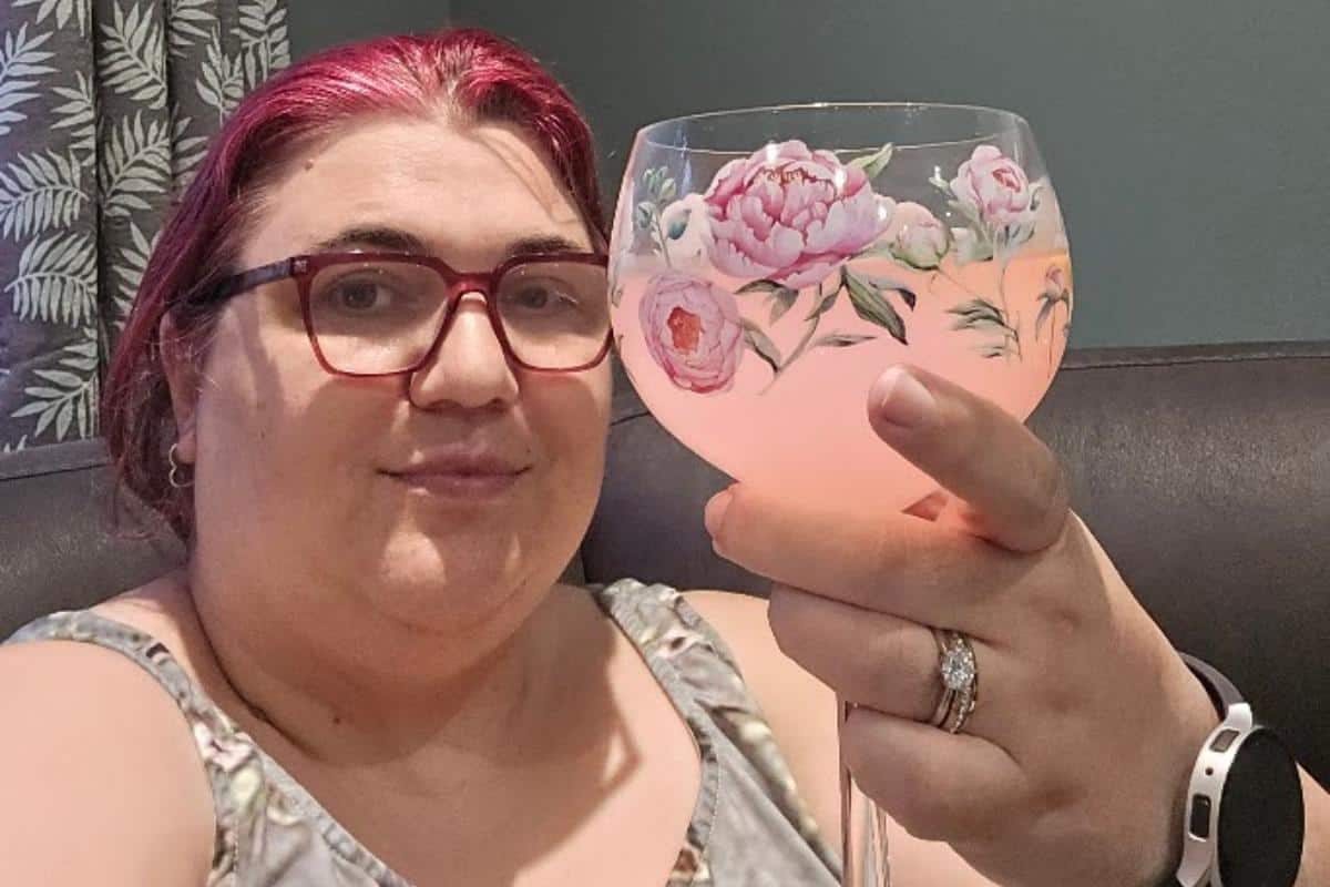 A person with pink hair and red glasses sits on a couch, holding up a floral-patterned glass filled with a pink gin for a taste test. They wear a sleeveless top, white smartwatch, and floral curtains hang in the background. Maybe it's Mr White's Gin!.