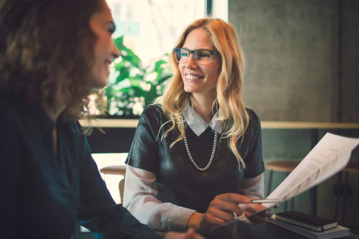 Two women sit at a table in an office, smiling and talking. One with long blonde hair and glasses holds papers, while the other listens. Sunlight shines through a window as they discuss returning to work after injury.