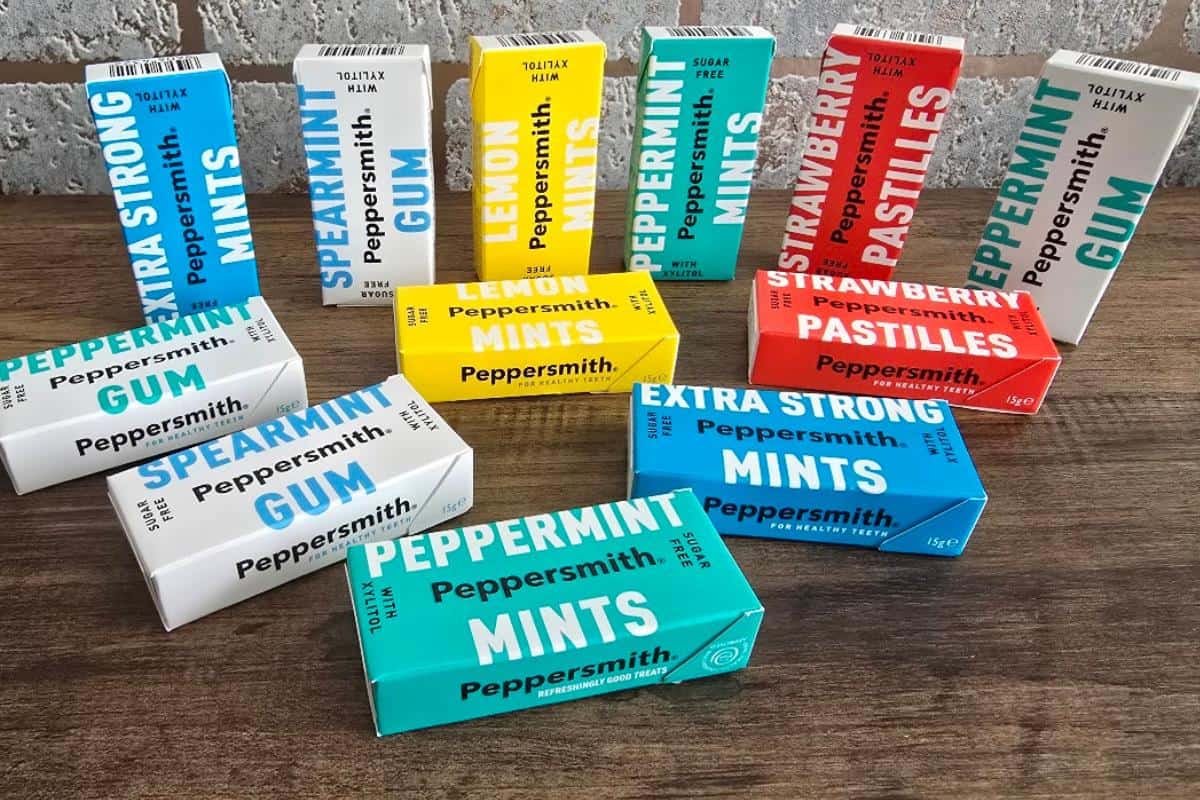 Various Peppersmith gum, mint, and pastille boxes in bright colors (blue, yellow, red, white) are arranged on a wooden table against a brick wall—a perfect addition to your European road trip essentials.