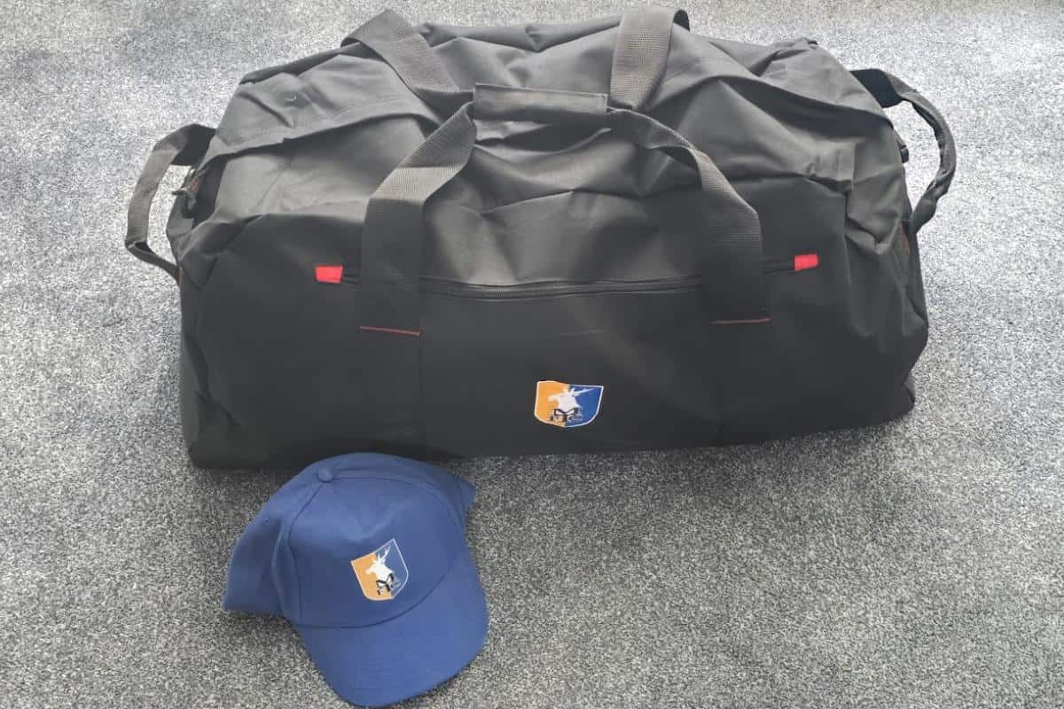 A large black duffel bag and a blue baseball cap, both featuring a logo with an orange and blue shield and black silhouette, are placed on a grey carpeted floor—perfect road trip essentials for your next European road trip.