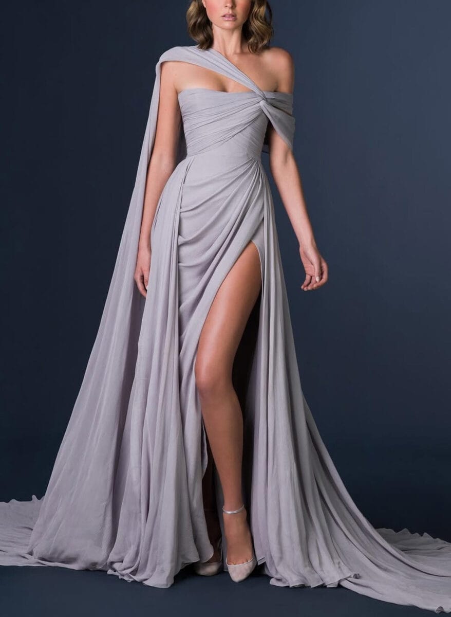 A woman stands in a flowing, pale lavender off-shoulder prom dress with a dramatic cape and high slit revealing one leg; the dress features draped, ruched fabric and pleats, paired with pointed ankle-strap heels against a dark background.