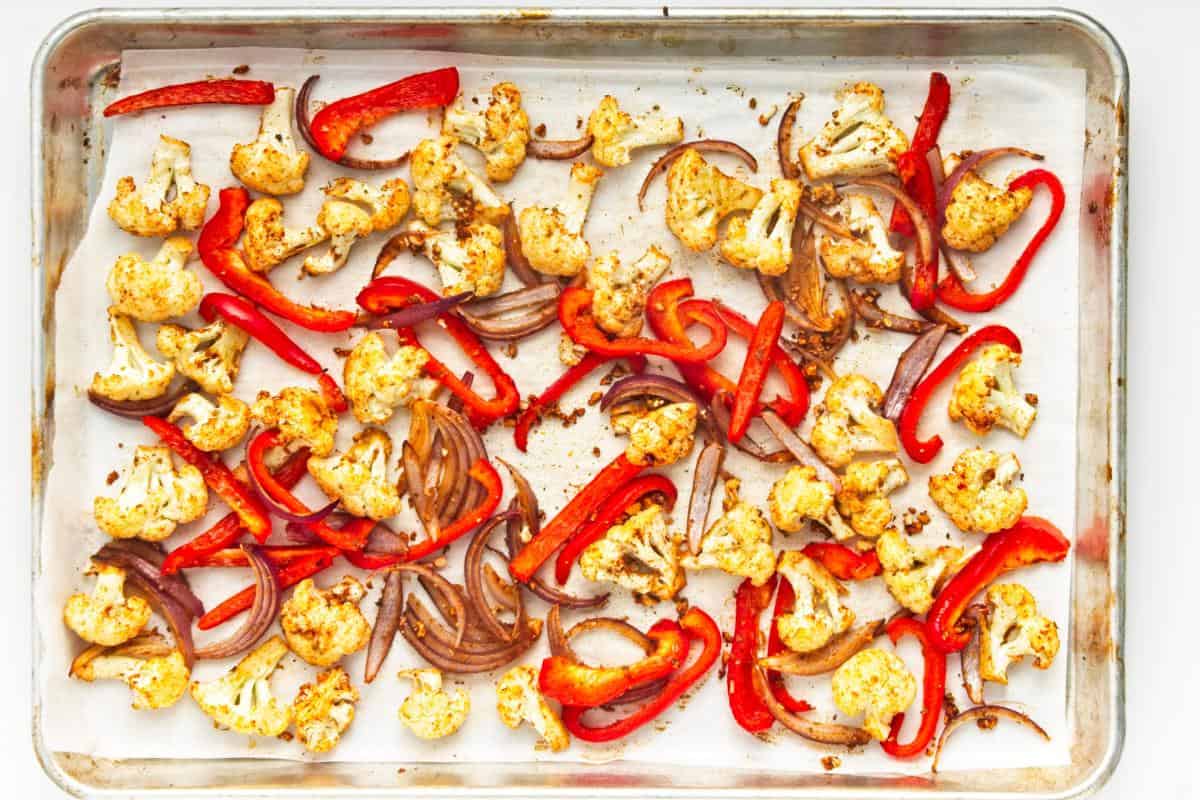 A baking sheet lined with parchment paper holds roasted cauliflower florets, red bell pepper strips, and sliced red onions—perfect for roasted cauliflower tacos or an easy dinner when paired with spicy cabbage slaw.