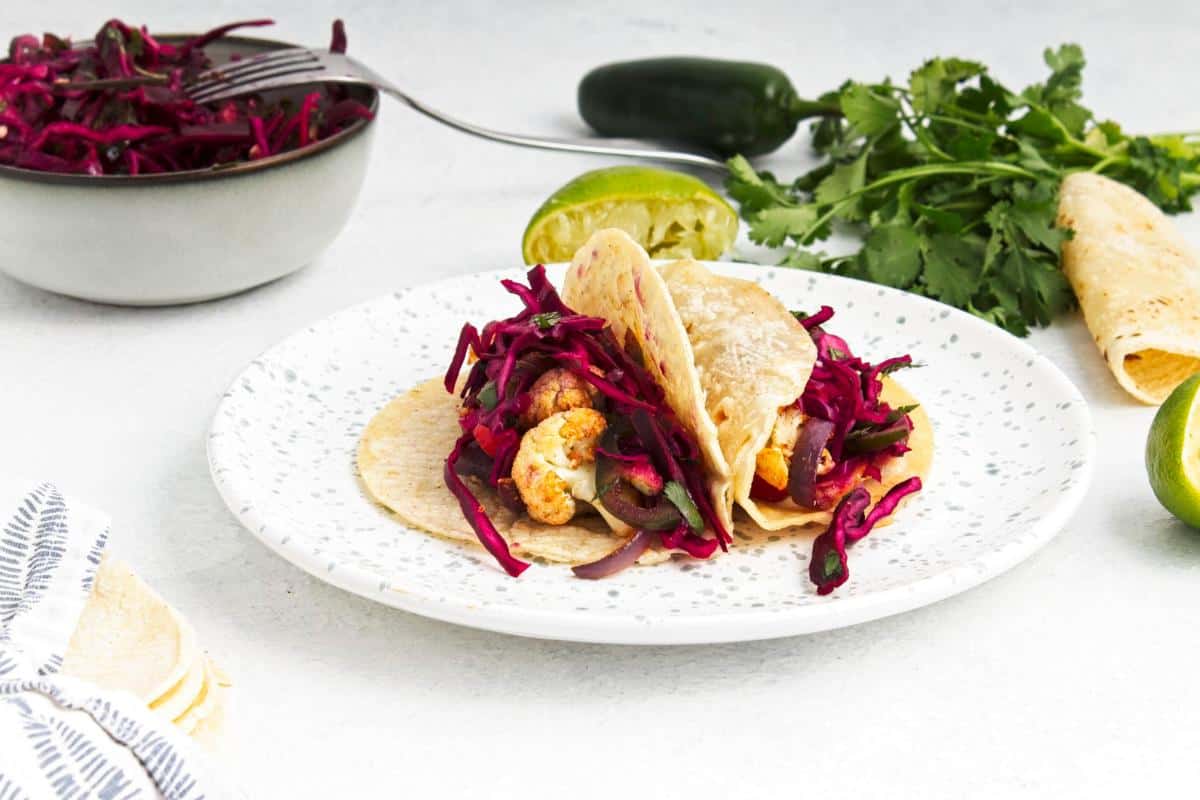 Two soft corn tortillas on a white speckled plate are filled with roasted vegetables and spicy cabbage slaw for easy Roasted Cauliflower Tacos. In the background: a halved lime, jalapeño, cilantro, folded tortillas, striped napkin, and a bowl of slaw.