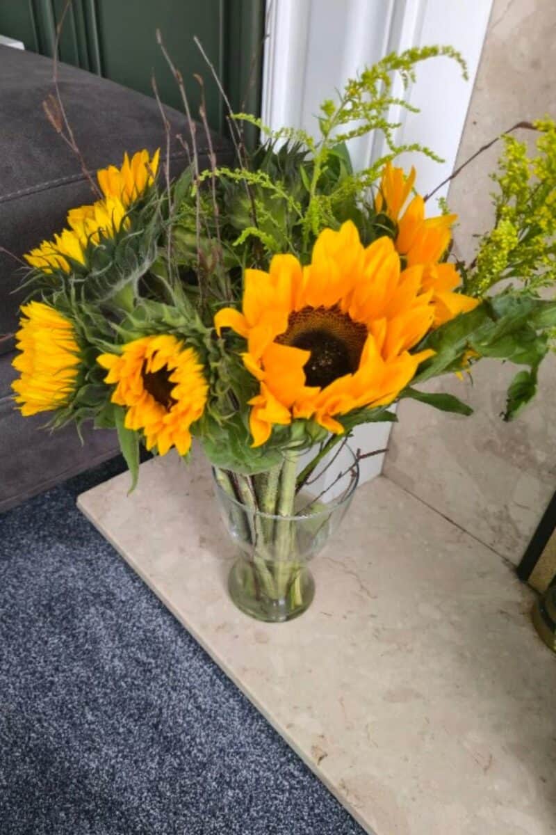 A clear glass vase holds a bouquet of bright yellow sunflowers from Prestige Flowers, with dark centers and green leaves. The tips have been followed to keep sunflowers fresh so these are vibrant and stunning.