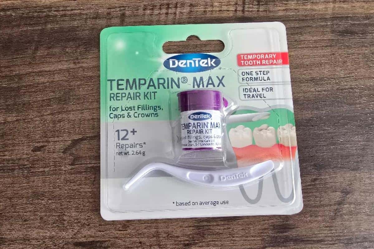 A DenTek Temparin Max repair kit for lost fillings, caps, and crowns sits on a wooden surface—an ideal road trip essential. The kit includes a small container and white applicator, with packaging highlighting “12+ repairs” for temporary tooth repair.