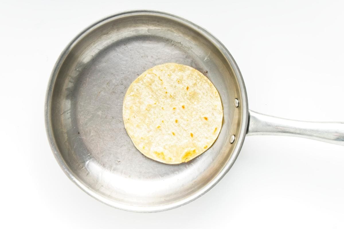 A single corn tortilla, perfect for Roasted Cauliflower Tacos or topped with Spicy Cabbage Slaw, heats in a stainless steel frying pan. The handle extends right, and the slightly browned tortilla is centered on the bright white background—an easy dinner idea.